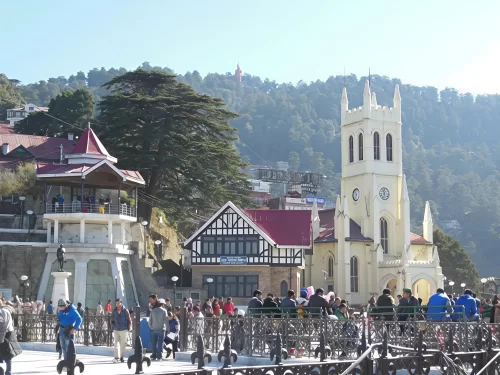 Crowded tourists at Christ Church The Ridge Shimla during sunny day, featuring Gothic tower half-timbered buildings statue fountain deodar hills Jakhoo Temple, perfect heritage cultural experience Himachal Pradesh tour packages.