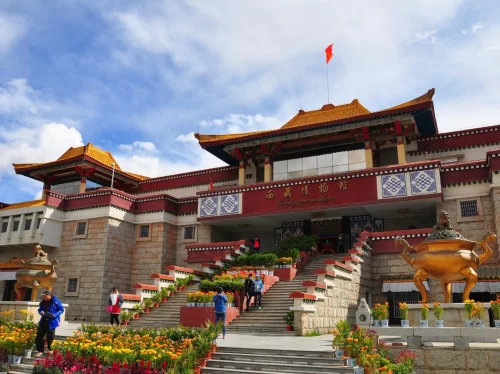 The Tibet Museum in Dharamshala featuring a grand Tibetan-style building with golden rooftops, red facade, wide staircase lined with flowers, and visitors at the entrance under a blue sky.