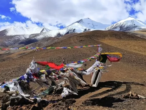 Thit Zarbo La Pass (Thang La) Ladakh Himalayas prayer flags fluttering amid brown rocky terrain with snow-capped peaks under blue sky with clouds, iconic high-altitude adventure trek tour package. 