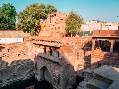 Toorji Ka Jhalra Bavdi stepwell at Jodhpur during golden hour, featuring red sandstone pavilions, arched gate, water pool, trees, perfect heritage experience Rajasthan tour packages.