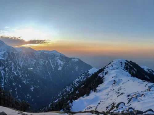Triund Dharamshala winter sunset panorama, featuring snow-covered Dhauladhar peaks golden orange skies trekking trails pine forests, perfect Himachal Pradesh adventure tour packages.