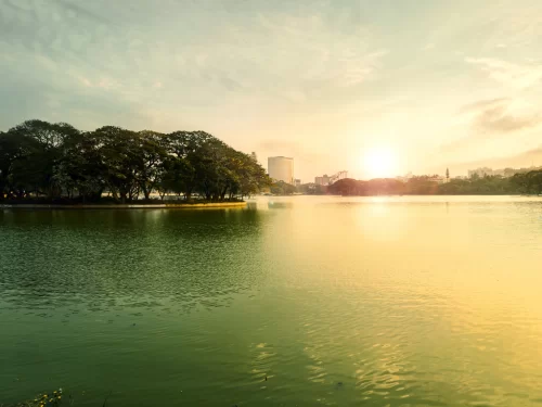Ulsoor Lake Bangalore golden hour sunset, lush trees lake highrises sun glow, perfect Karnataka tour package.