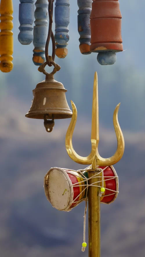 Sacred trishul with damru and hanging temple bells set against a serene Himalayan backdrop, symbolizing the spiritual essence of Char Dham in Uttarakhand, a highlight of Uttarakhand tour packages.