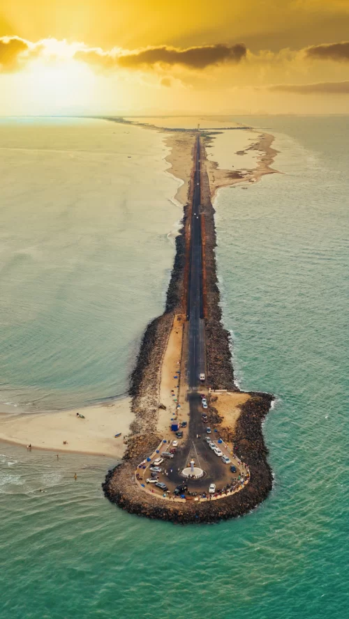 Aerial view of Dhanushkodi road stretching into the sea at Rameswaram, Tamil Nadu, with golden sunset skies and turquoise waters, a breathtaking coastal attraction featured in Tamil Nadu tour packages.