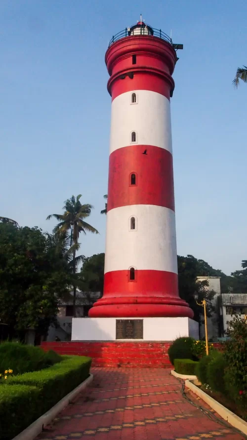 Red and white lighthouse standing tall in Alleppey, Kerala, surrounded by palm trees and manicured gardens, a charming coastal landmark featured in Kerala tour packages.