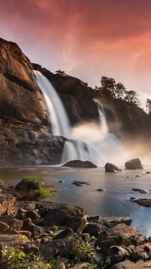 Scenic Athirappilly Waterfalls flowing over rocky cliffs at sunset in Kerala, surrounded by mist and lush greenery, a stunning natural attraction featured in Kerala tour packages.