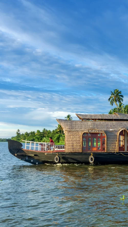 Traditional Kerala houseboat sailing through the scenic backwaters of Kumarakom, surrounded by palm trees and calm blue waters under a clear sky.