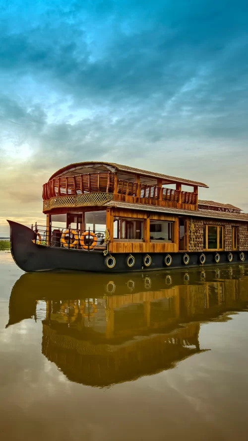 Luxury houseboat floating on the tranquil backwaters of Kumarakom, Kerala, with wooden interiors and its reflection shimmering on calm waters at sunset.