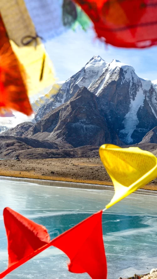 Colorful prayer flags fluttering near Gurudongmar Lake in Sikkim with snow covered Himalayan peaks and frozen turquoise waters featured in Sikkim tour packages