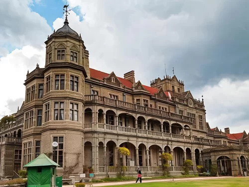 Viceregal Lodge Shimla grand colonial architecture during partly cloudy day, featuring red roofs arched verandas manicured lawns person walking, perfect heritage romantic experience Shimla tour packages.