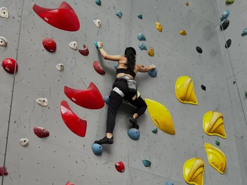 Wall Climbing activity with a woman climbing an indoor artificial rock wall, gripping colorful holds while wearing climbing shoes and safety gear in a modern climbing gym.