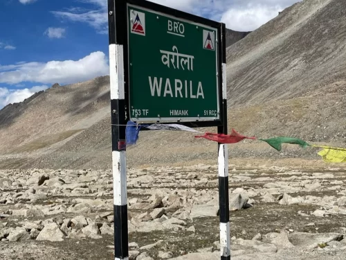Wari La Pass Ladakh official BRO green signboard marking high-altitude pass under Himank Project with colorful Tibetan prayer flags against barren rocky Himalayan mountains, offbeat Nubra Valley route