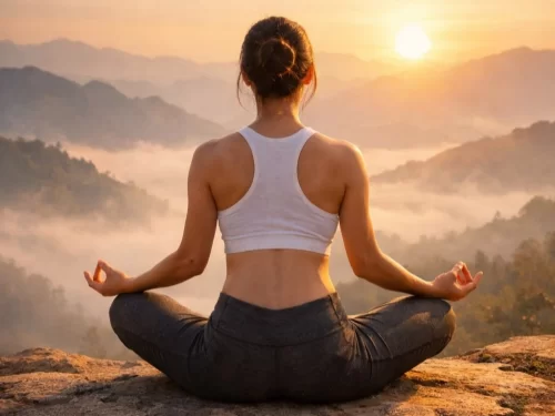 A woman practicing yoga and meditation in a cross-legged pose on a hilltop, facing a serene sunrise over mist-covered mountains, creating a peaceful and calming atmosphere.