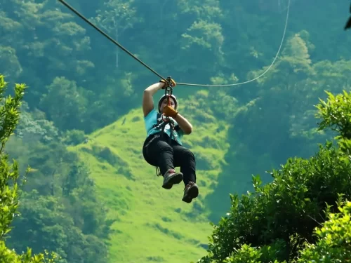 Ziplining adventure showing a person suspended from a cable, gliding above lush green hills while secured with a safety harness and pulley system, surrounded by scenic forested mountains.