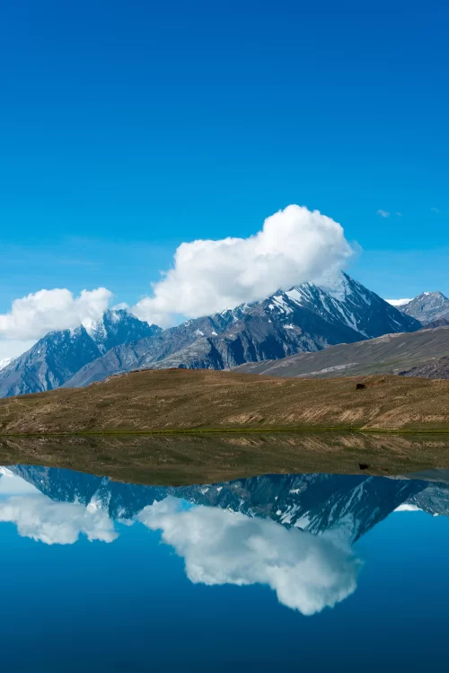 Crystal clear reflections of snow capped peaks in Chandra Tal Lake under bright blue sky, a breathtaking high altitude wonder featured in Himachal Pradesh tour packages.