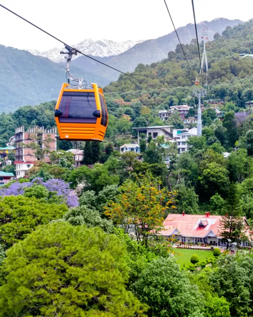 Orange cable car gliding above the scenic hills of Dharamshala, Himachal Pradesh, with lush green forests, hillside homes, and snow-capped mountains in the background.