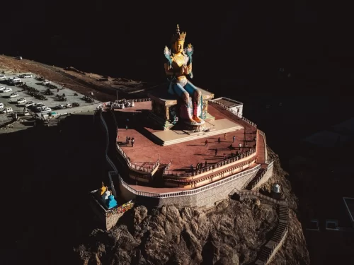 Diskit Gompa Ladakh aerial drone view golden Maitreya Buddha statue hilltop red platform parking cars rock cliff Nubra Valley, perfect Leh Ladakh spiritual adventure package.