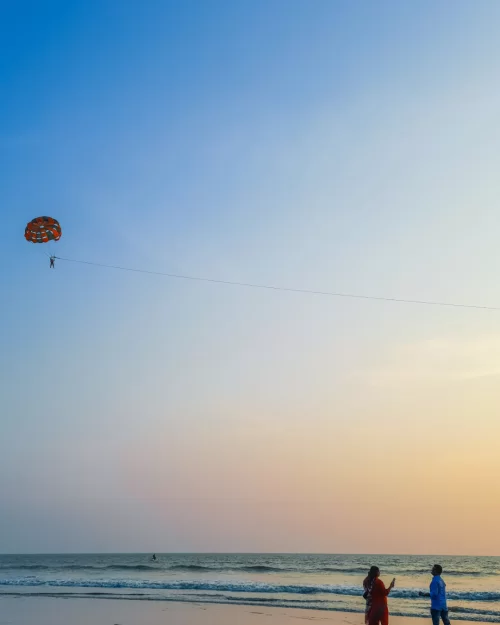 Parasailing adventure over the Arabian Sea at sunset with visitors enjoying the beach in Goa, an exciting activity featured in Goa tour packages.