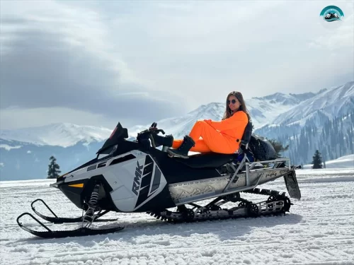 Gulmarg Kashmir snow slope orange suit woman black white snowmobile sunglasses snowy mountains cloudy skies pine trees, perfect winter snowmobile adventure tour package. 