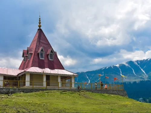 Gulmarg Kashmir maroon temple conical roof golden pinnacle flags tourists yellow fence snowy mountains pine forests green grass blue cloudy skies, perfect Shiva shrine Himalayan spiritual tour package.
