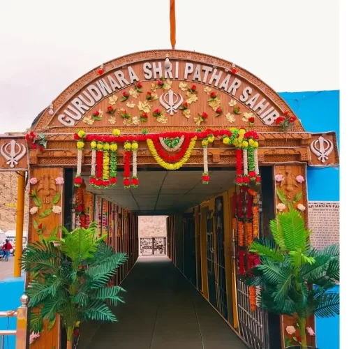 Ornate duplicate entrance archway at Gurdwara Shri Pathar Sahib Leh Ladakh showcasing carved wooden gateway saffron flag floral marigold garlands Khanda emblems green plants corridor blue walls rocky terrain vehicles, colorful inviting spiritual Ladakh to