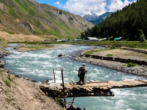 Elder crossing wooden bridge at Gurez Valley during sunny partly cloudy weather, featuring turquoise river and green mountains, perfect adventure Srinagar tour package. 