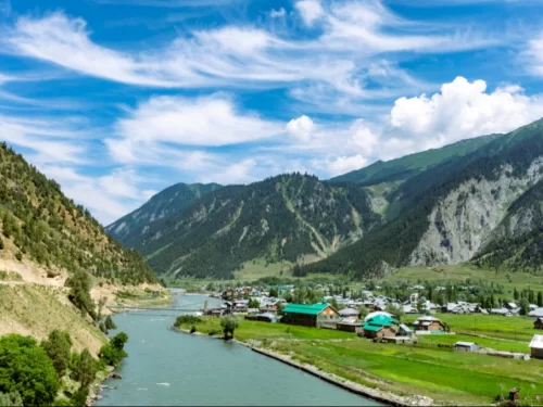 Village panorama at Gurez Valley during sunny partly cloudy weather, featuring green fields and turquoise river, perfect adventure Srinagar tour package.