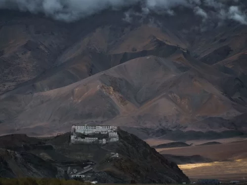 Hanle Gompa perched on hill amid dramatic Ladakh mountains during cloudy dusk, featuring white buildings against barren slopes, perfect Ladakh tour package.