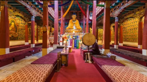 Interior of Dukhang main prayer hall Hemis Gompa Himis Monastery Ladakh India featuring large golden Shakyamuni Buddha statue colorful wall murals red pillars maroon benches monk playing drum prayer flags hanging, serene Tibetan Buddhist gompa ambiance