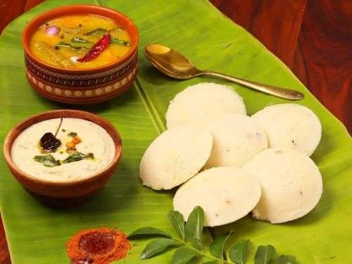 Idli sambar features soft steamed rice cakes with spicy lentil vegetable stew, coconut chutney and red chili powder on banana leaf, authentic South Indian breakfast delight.
