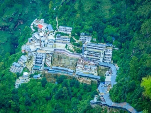 Aerial view Vaishno Devi Bhawan Katra Jammu Kashmir daytime, featuring white red roofed buildings perched forested Trikuta mountains green slopes stairs backdrop, perfect pilgrimage experience Jammu Kashmir tour packages.
