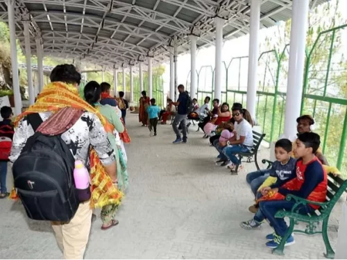 Pilgrims families waiting Katra railway station Jammu Kashmir daytime, featuring saffron dupattas backpacks benches green railing hills backdrop Vaishno Devi yatra, perfect pilgrimage experience Jammu Kashmir tour packages.