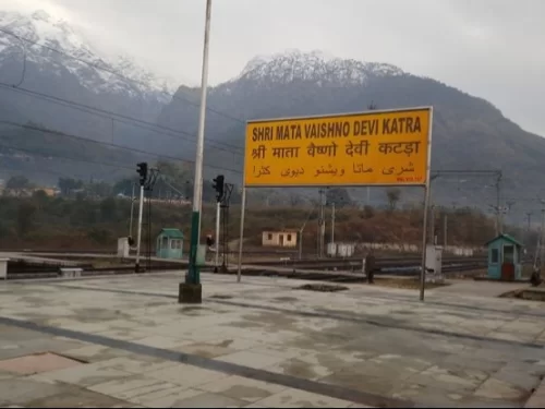 Shri Mata Vaishno Devi Katra railway station Jammu Kashmir cloudy day, featuring yellow signboard snowy Trikuta mountains signals tracks backdrop pilgrimage base, perfect pilgrimage experience Jammu Kashmir tour packages.