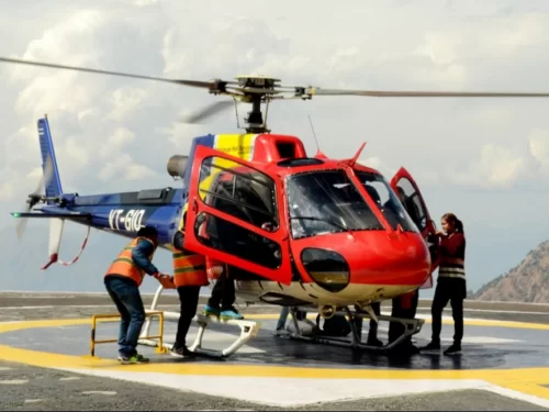 Red blue helicopter VT-GJD helipad Katra Jammu Kashmir sunny day, featuring ground crew loading doors open Trikuta mountains backdrop Vaishno Devi helicopter service, perfect pilgrimage experience Jammu Kashmir tour packages.