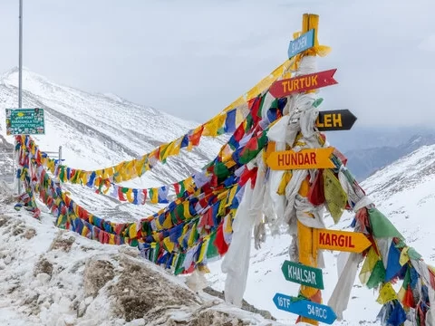 Colorful prayer flags and signposts pointing to Turtuk, Leh, Hundar, Kargil and Khalsar at snow-covered Khardungla Pass, capturing high-altitude Himalayan adventure vibes.