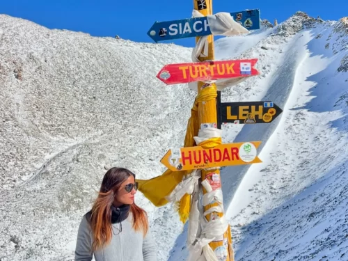 Signpost at Khardungla Pass during snowy daytime, featuring colorful signs to Siachen Diskit Leh, prayer flags, rugged peaks, perfect adventure experience with Ladakh tour package.