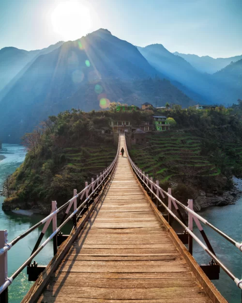 Wooden suspension bridge leading toward a small hillside village in Kullu, Himachal Pradesh, with river waters below and sun rays streaming over the surrounding Himalayan mountains.