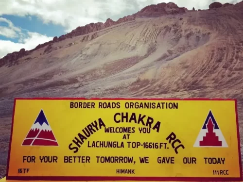 Bright yellow BRO board at Lachungla Top (16,616 ft) with Shaurya Chakra RCC welcomes your message, set against barren brown mountains and a partly cloudy high-altitude sky.