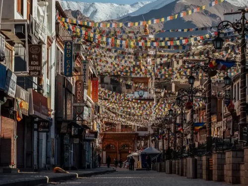 Early morning view of Leh Market with Tibetan prayer flags hanging over quiet street, lined shops and cafes leading towards snow-capped mountains, perfect for Leh city walking tour blogs