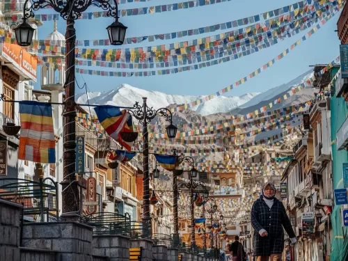 Daytime scene of Leh Market with a lone walker, rows of shops and ornate lamp posts draped in colourful prayer flags, framed by snow-covered Himalayan peaks, ideal for Leh city street photography blogs
