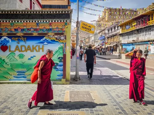 Vibrant Leh Market street scene with monks in maroon robes, colourful “I Love Ladakh” wall art, prayer flags and bustling shops under clear blue sky, ideal for Ladakh culture and street photography blogs