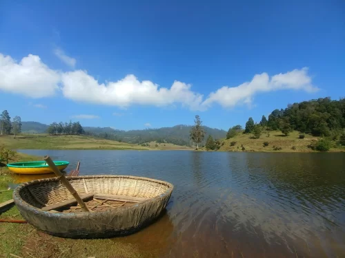 Mannavanur Lake near Kodaikanal, featuring coracle boat on calm blue waters, rolling green hills and clear sky, perfect nature Tamil Nadu tour package