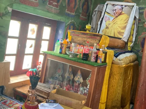 Cozy Mulbekh Monastery prayer altar shrine with Dalai Lama portrait colorful Tibetan thangka murals glass-cased figurines offerings bottles snacks incense yellow silk cushions green rug wooden cabinets under soft indoor light Ladakh, perfect India Leh Kar