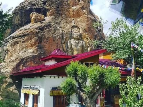 Towering 9-meter rock-carved Maitreya Buddha Future Buddha statue atop Mulbekh Gompa monastery with red-tiled roof white walls fluttering prayer flags lush green trees rugged brown cliffs cloudy skies Ladakh India, perfect Leh Kargil Himalayas Buddhist ro