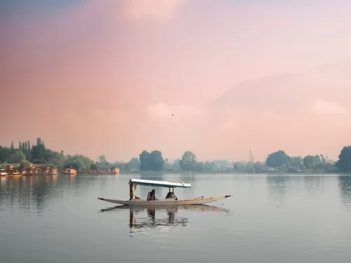 Shikara ride on Nigeen Lake in Srinagar at sunset, with houseboat passengers, misty pink skies, Zabarwan mountains backdrop, serene Kashmir houseboat experience.