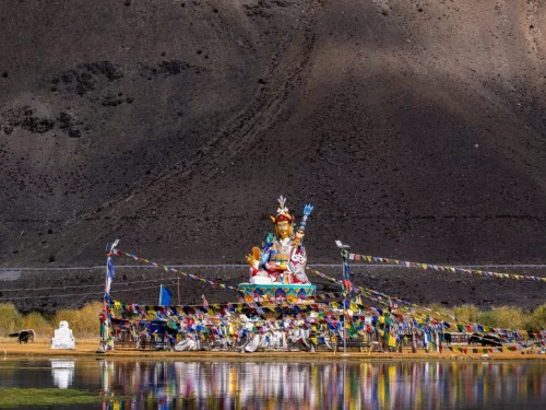 Guru Padmasambhava statue at Sani Lake near Padum village Zanskar Valley Ladakh during clear day, featuring prayer flags, motorcycles, barren mountain backdrop, perfect spiritual Zanskar tour package. 