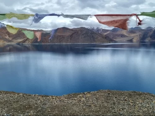 Ladakh Pangong Tso Lake turquoise blue brackish water prayer flags multicolored Tibetan Buddhist fluttering rocky shore snow capped rugged brown Himalayan mountains cloudy sky high altitude pilgrimage adventure tour package. 
