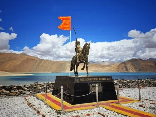 Ladakh Pangong Lake Chhatrapati Shivaji Maharaj golden bronze equestrian statue saffron flag pedestal black base railing yellow markings turquoise blue lake brown barren mountains blue sky historical monument adventure tour package. 