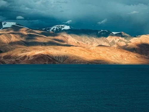 Ladakh Pangong Tso Lake dramatic sunset golden orange light snow capped rugged brown Himalayan mountains dark blue brackish water cloudy sky high altitude scenic landscape adventure tour package Ladakh Himalayas.