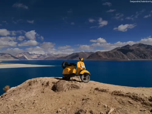 Ladakh Pangong Tso Lake iconic yellow Vespa scooter sidecar on rocky cliff overlooking turquoise blue high altitude lake snow capped Himalayan mountains barren brown landscape blue sky adventure tour package.